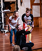 Jenny Bennett sitting on the front of the stage and wearing black shirt and pants holds up the play program  for Ginna Hoben in white t-shirt and geans, and Allie Babich in black and gray t-shirt ad red and yellow tights, both leaning over to read what Bennett is pointing at in the program